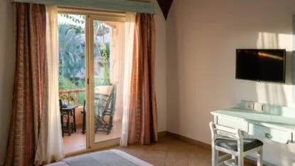 Camera da letto con scrivania, sedia, TV a parete e balcone con mobili da esterno e vegetazione.