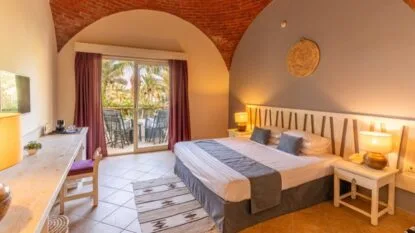 Camera da letto accogliente con letto matrimoniale, parete con archi in mattoni, scrivania e balcone con vista su palme.
