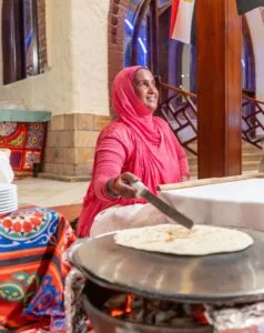 Donna sorridente con abito rosa mentre cucina pane tradizionale su piastra calda in ambiente interno arabo.