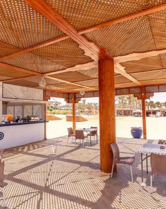 Veranda ombreggiata con travi di legno intagliato, tavoli e sedie su spiaggia soleggiata con palme sullo sfondo.