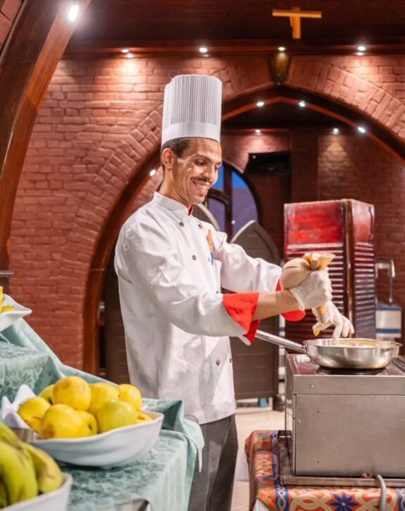 Chef sorridente in uniforme bianca che cucina in una cucina rustica con archi in mattoni rossi.