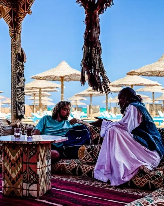 Due uomini seduti su divani tradizionali decorati, conversano in un lounge all'aperto con ombrelloni di paglia sullo sfondo.