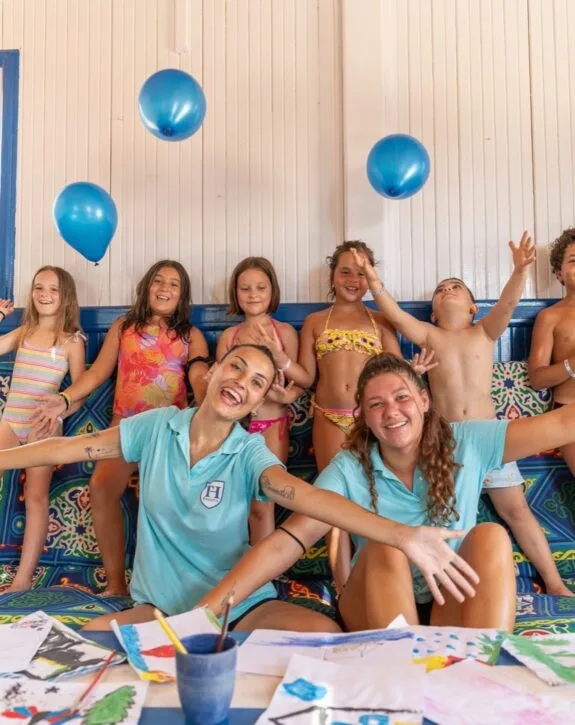 Bambini in costume con palloncini blu e due ragazze sorridenti con polo azzurre sedute davanti a disegni colorati.