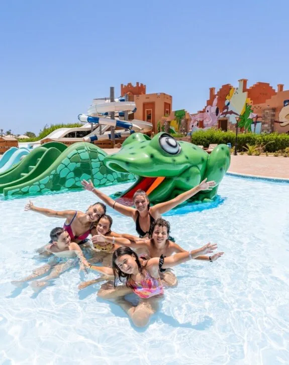 Bambini sorridenti che giocano in piscina davanti a uno scivolo a forma di rana verde in un parco acquatico.