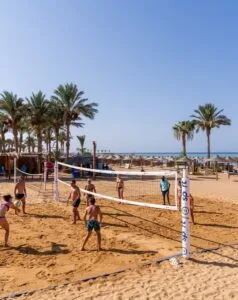 Persone che giocano a beach volley su spiaggia sabbiosa con palme e ombrelloni di paglia sullo sfondo.
