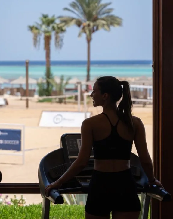 Donna in abbigliamento sportivo sul tapis roulant con vista sulla spiaggia, palme e mare al tramonto.