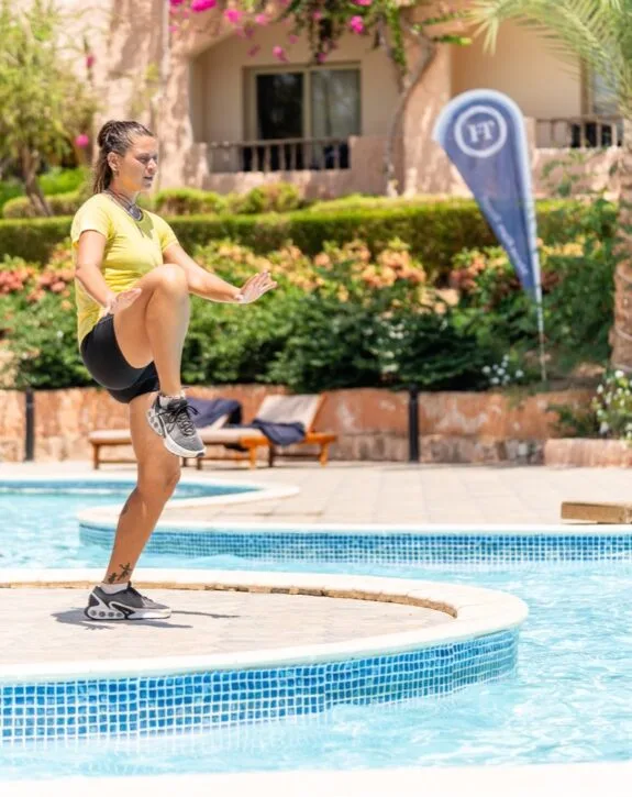Donna che si esercita in equilibrio su una gamba vicino a una piscina in un ambiente esterno soleggiato.