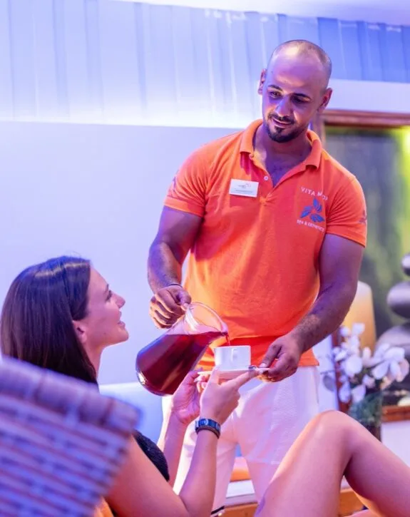Operatore in polo arancione versa bevanda in tazza mentre donna seduta sorride in ambiente rilassante spa.