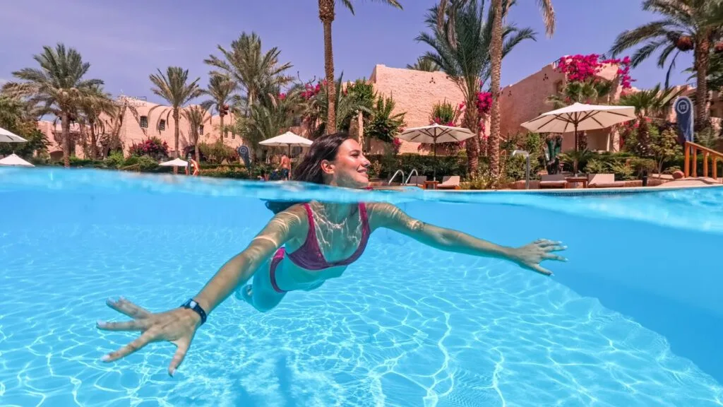 Donna che nuota sott'acqua in piscina con sfondo di palme e ombrelloni in un resort soleggiato.