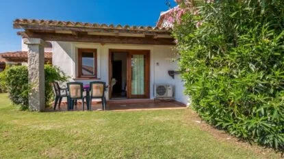 Casa con veranda, tavolo e sedie, circondata da verde e fiori, sotto un cielo sereno.
