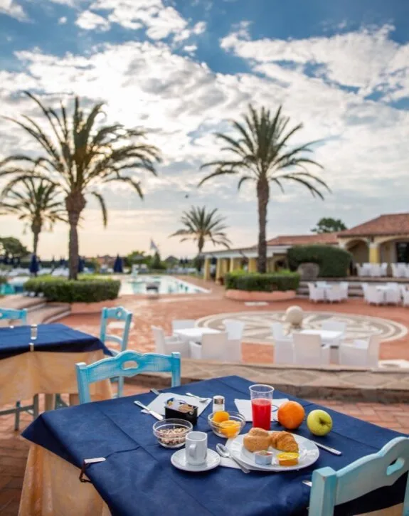 Colazione all'aperto con tavolo, frutta, bevande e piscina sullo sfondo in un resort con palme e cielo nuvoloso.