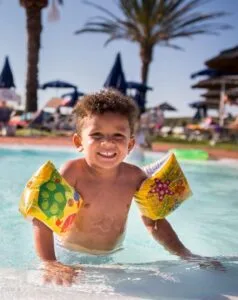 Bambino sorridente in acqua con braccioli colorati, circondato da palme e sdraio al sole.