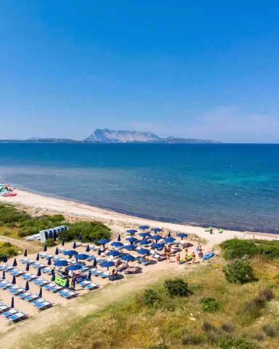 Spiaggia affollata con ombrelloni blu, lettini e vista sul mare turchese e un'isola sullo sfondo. Atmosfera estiva.