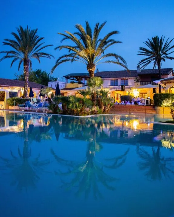 Vista serale di una piscina con palme, riflessi nell'acqua e ambiente rilassato attorno a un ristorante.