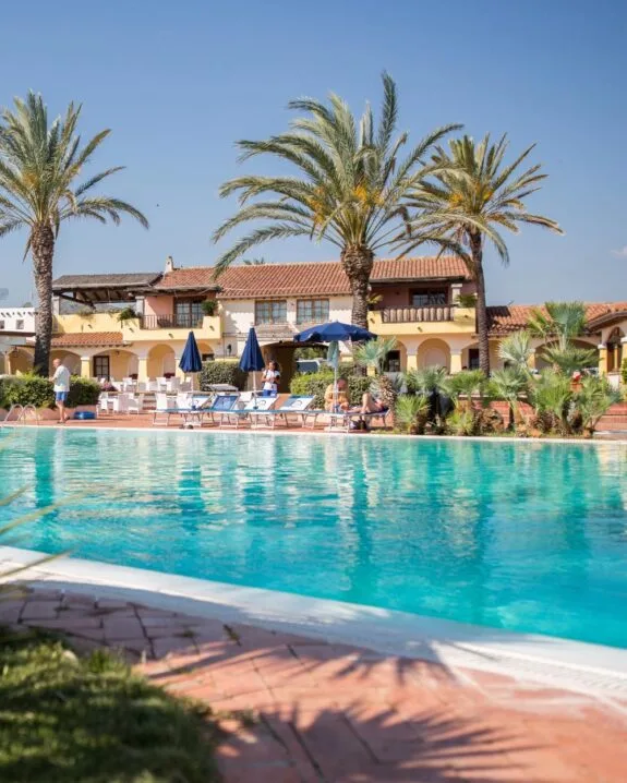 Piscina azzurra circondata da palme, lettini e edifici in stile mediterraneo, sotto un cielo sereno.