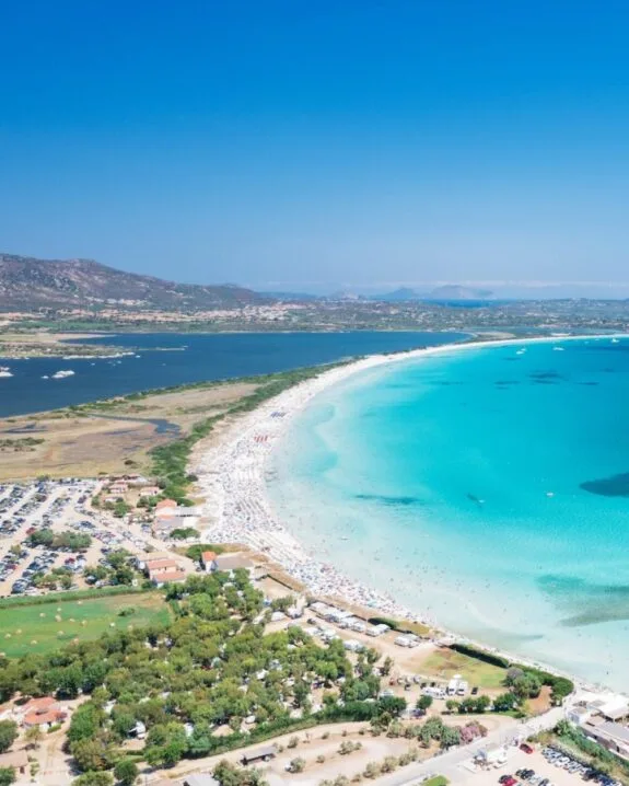 Spiaggia con sabbia bianca e acqua turchese, affollata di persone e ombrelloni, circondata da colline verdi.