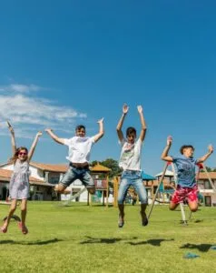 Bambini che saltano in un parco giochi all'aperto con case e giochi sullo sfondo, cielo azzurro e giornate di sole.