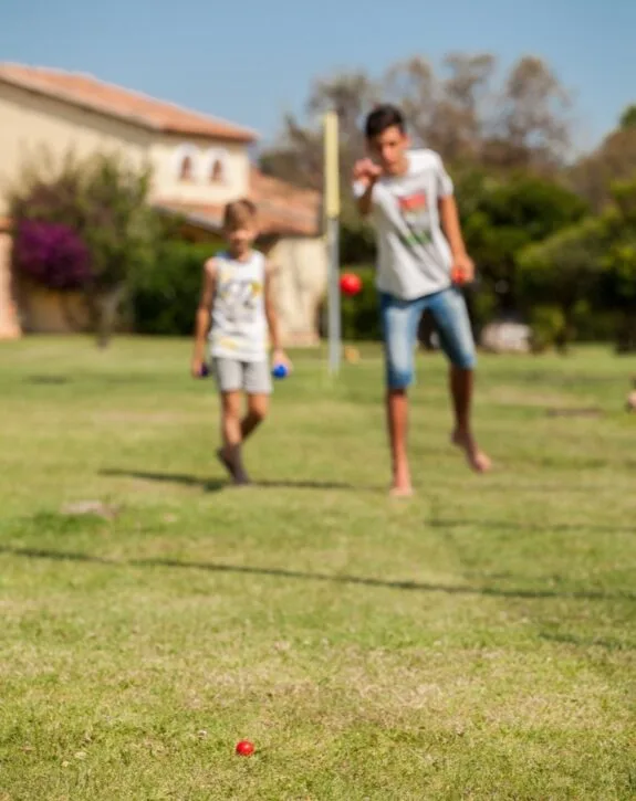 Due bambini giocano con bocce su un prato soleggiato, uno mira verso la palla rossa.