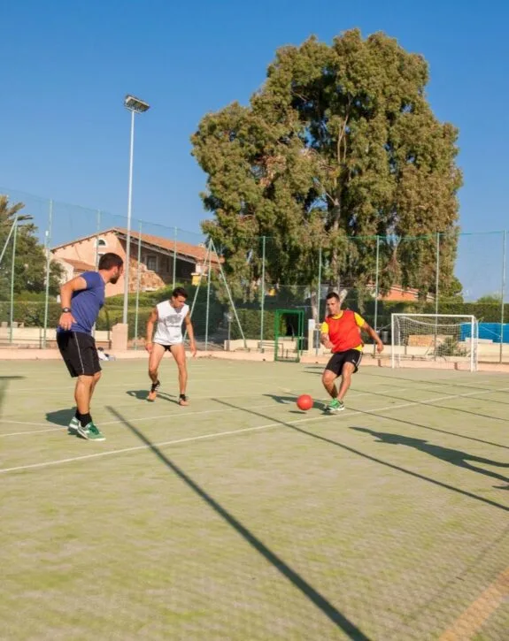 Tre persone che giocano a calcio su un campo all'aperto con rete e albero grande sullo sfondo.