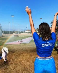Donna con maglietta e pantaloncini azul, che si prepara a scivolare sul prato con un thumbnail bagnato, mentre un arcobaleno si forma dall'acqua.