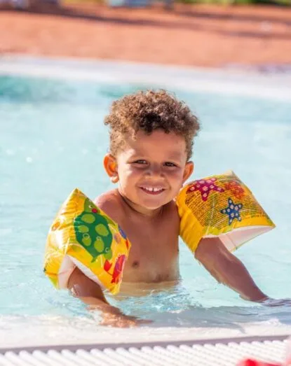 Bambino sorridente con braccia gonfiabili in piscina, acqua azzurra e bordo in primo piano.