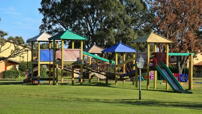 Parco giochi con casette colorate e scivoli, alberi sullo sfondo e prato verde.