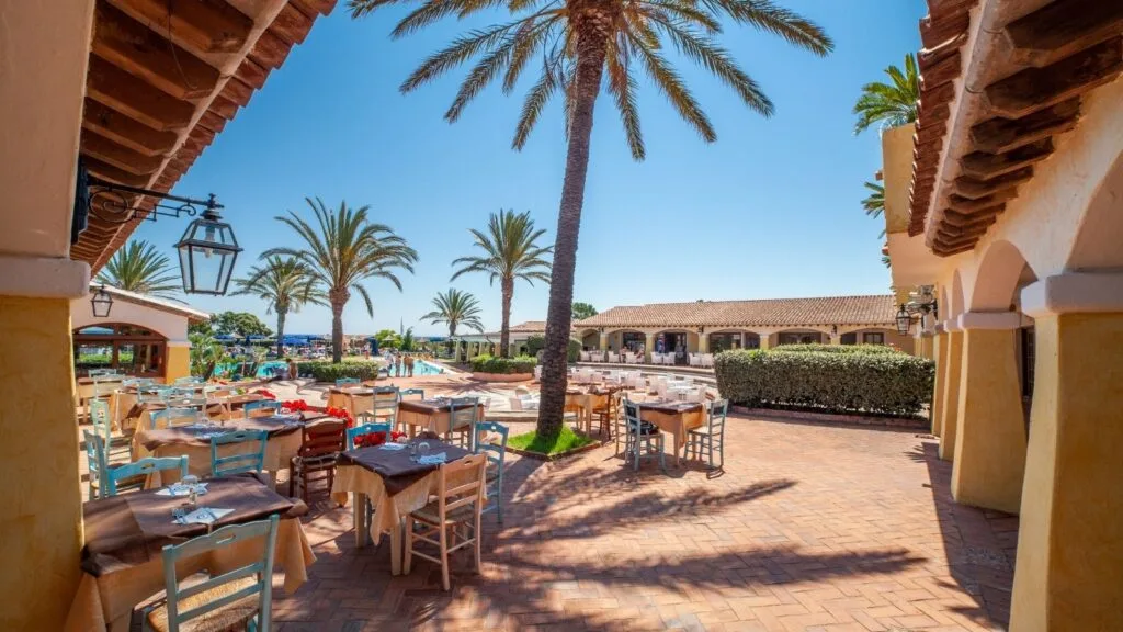Ristorante all'aperto con tavoli, palme e piscina sotto un cielo blu soleggiato.