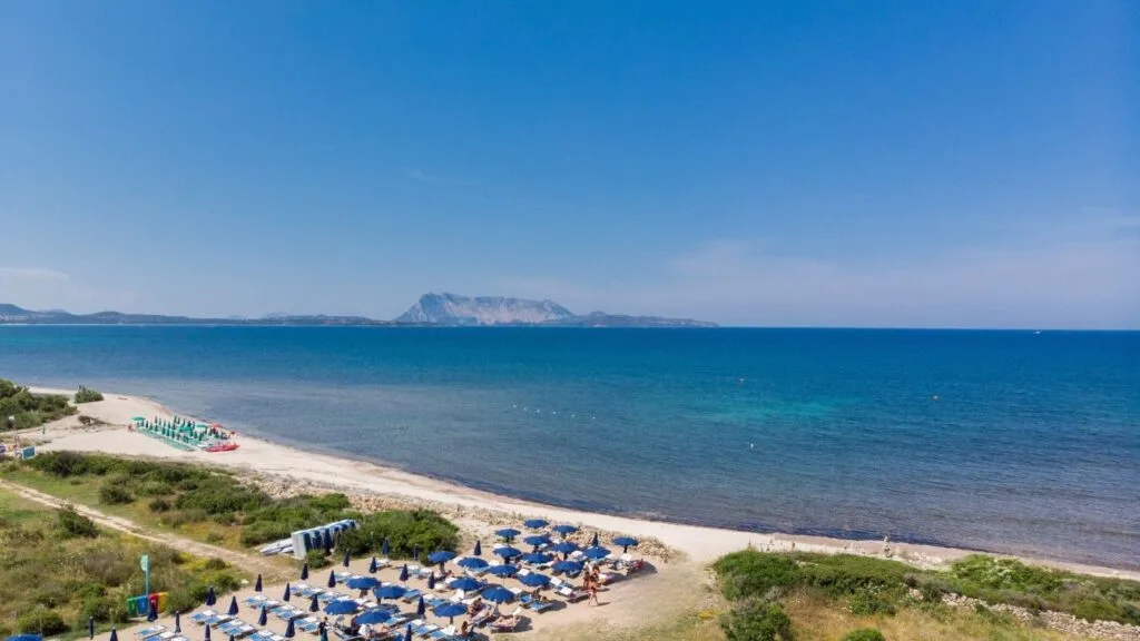 Spiaggia con ombrelloni e lettini, mare e cielo azzurro con isola sullo sfondo.
