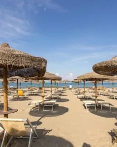 Spiaggia sabbiosa con lettini e ombrelloni in paglia, oltre una distesa di mare sotto un cielo sereno e azzurro.