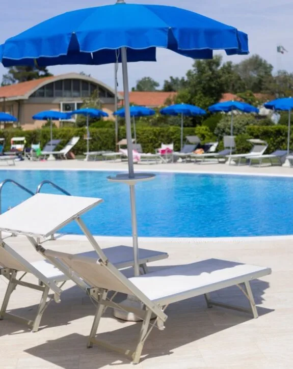 Lettini bianchi con ombrelloni blu vicino a una piscina in una giornata soleggiata.
