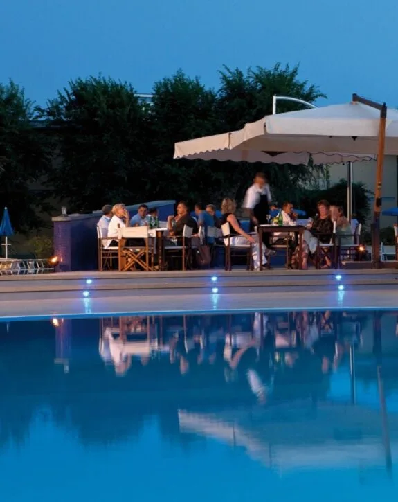 Persone che cenano all'aperto vicino a una piscina illuminata di sera sotto un ombrellone grande.