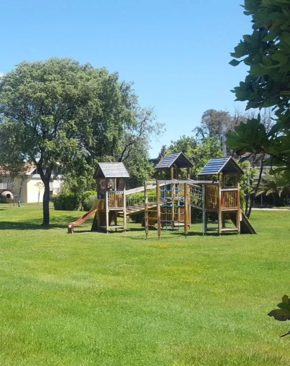 Parco giochi in legno con scivolo e arrampicata su prato verde, alberi e cielo sereno sullo sfondo.