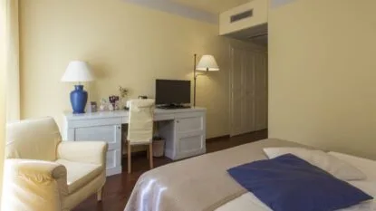 Camera da letto con letto, scrivania con computer, poltrona beige e lampade su pareti gialle.