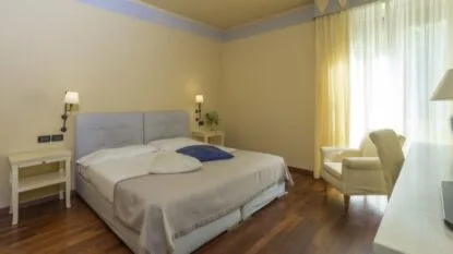 Camera da letto luminosa con letto matrimoniale, comodini, poltrona bianca e tende gialle in ambiente moderno.