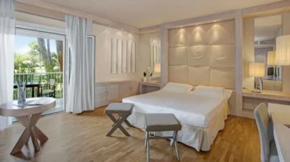 Camera da letto moderna con letto matrimoniale, due sgabelli, tavolo rotondo, lampade e finestra con vista sul verde.