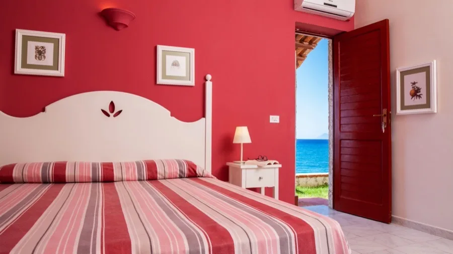 Camera con parete rossa, letto con copriletto a righe rosse, portafinestra aperta con vista sul mare azzurro.