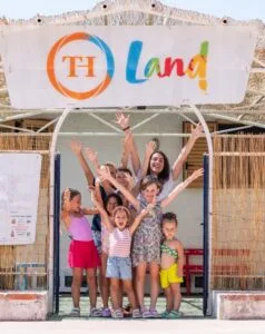 Gruppo di bambini sorridenti e una donna con le mani alzate sotto un arco con insegna colorata "TH Land!"