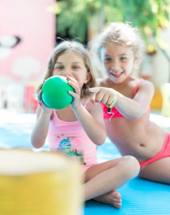 Due bambine sorridenti sedute su un tappetino, una tiene un palloncino verde e l'altra indica davanti a sé.
