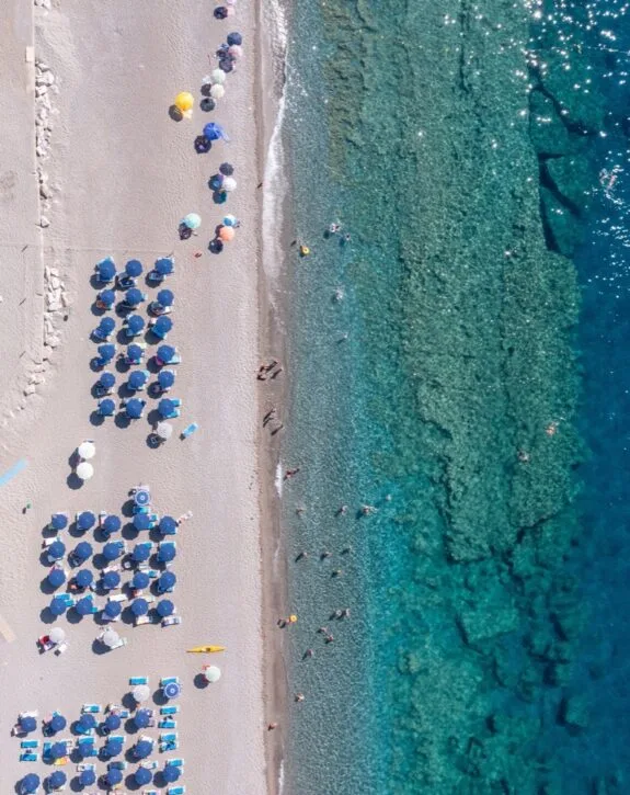 Spiaggia con ombrelloni blu e persone che nuotano nel mare cristallino visto dall'alto.