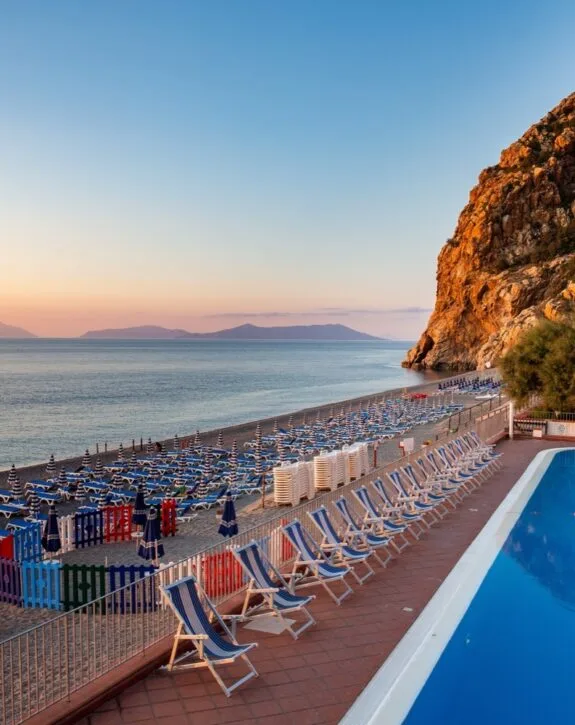 Spiaggia con lettini e ombrelloni blu, piscina e grande scogliera al tramonto sul mare calmo.