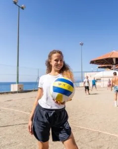 Ragazza sorridente con pallone da beach volley in mano su campo sabbioso vicino al mare, cielo limpido.