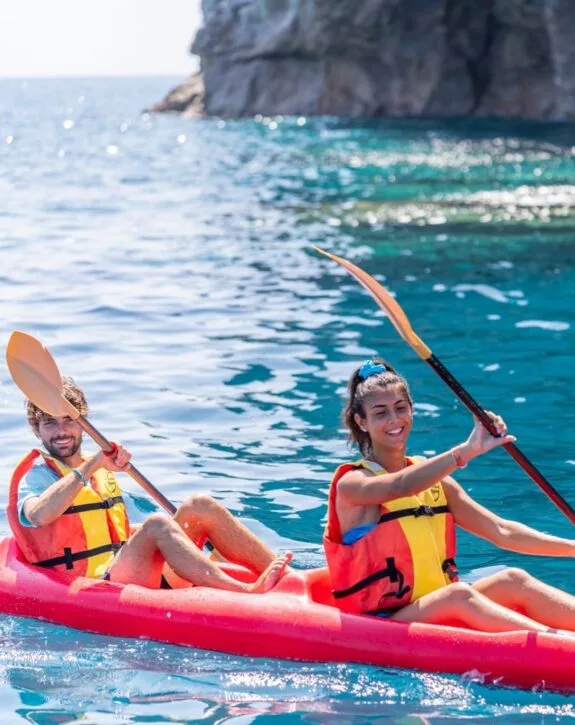 Due persone con giubbotti di salvataggio rossi e gialli che fanno kayak su acqua blu vicino a una scogliera.