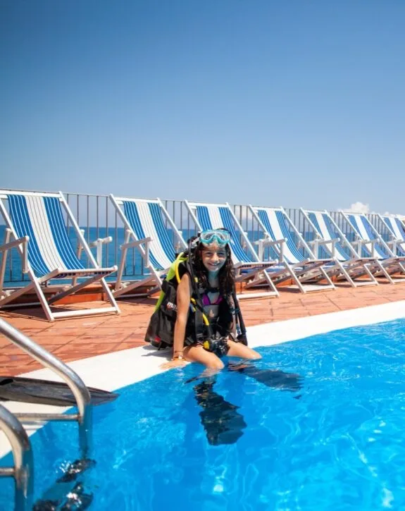 Ragazza con attrezzatura da sub seduta sul bordo di una piscina con sedie a sdraio blu e bianche sullo sfondo.