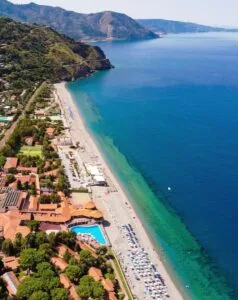 Spiaggia attrezzata con ombrelloni, mare turchese e edifici con piscina vicino a colline verdi sullo sfondo.