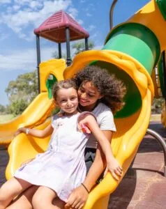 Due bambine sorridenti scendono insieme uno scivolo giallo in un parco giochi soleggiato.