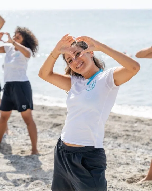 Ragazza sorridente con maglietta bianca che forma un cuore con le mani sulla spiaggia, mare sullo sfondo.