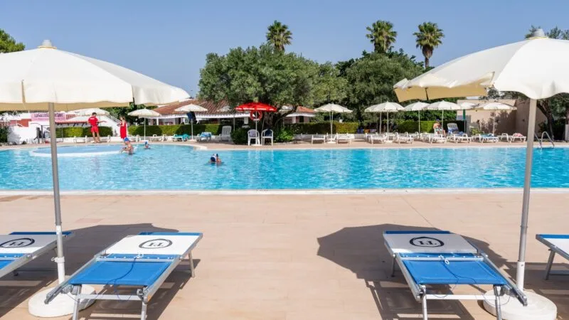 Piscina all'aperto con sdraio blu, ombrelloni bianchi e persone che nuotano in una giornata soleggiata.