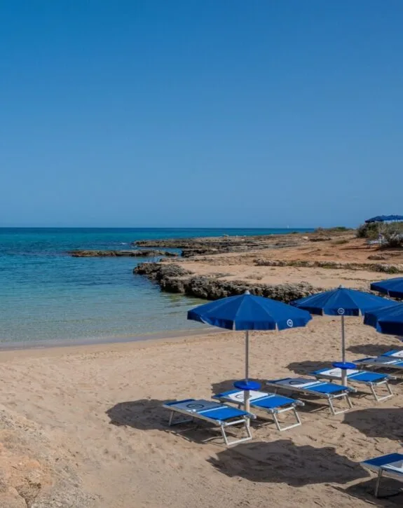 Spiaggia con sabbia chiara, ombrelloni blu e lettini, mare blu e roccia a sinistra, cielo sereno senza nuvole.