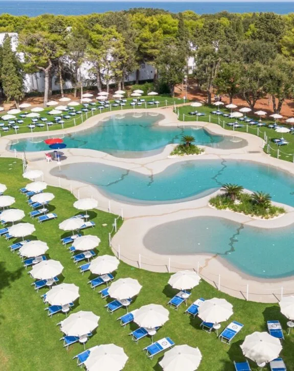 Piscina a forma organica circondata da lettini e ombrelloni bianchi, con vista su alberi verdi e mare in lontananza.