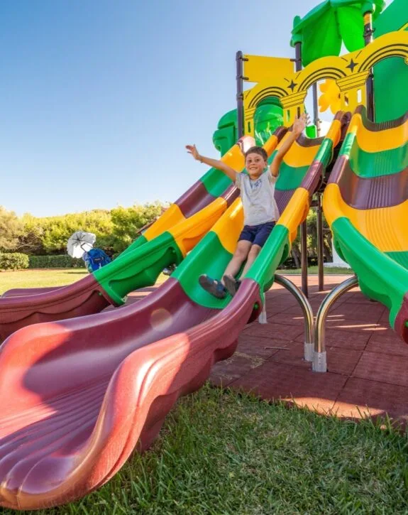 Bambino felice che scende da uno scivolo colorato a tre corsie in un parco giochi all'aperto.